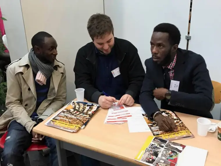 Trois hommes discutent autour d'une table couverte de brochures d'information.