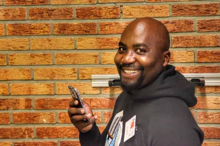Un homme souriant devant un mur de briques, son téléphone à la main.